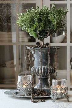 Black urn with a boxwood topiary planted in it and chains hanging down on dining table