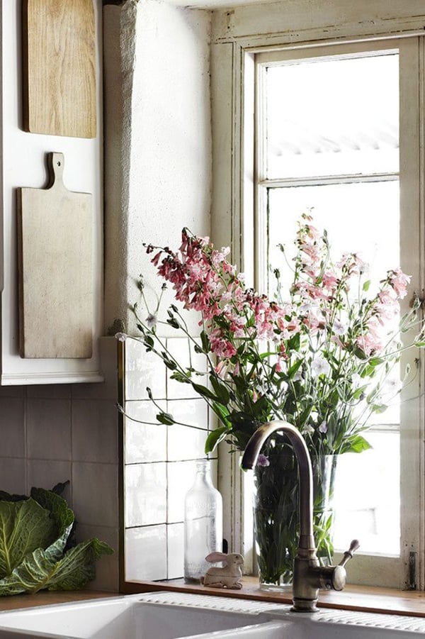 Rustic kitchen with tall arrangement of pink flowers in window behind sink and vintage bread boards on wall