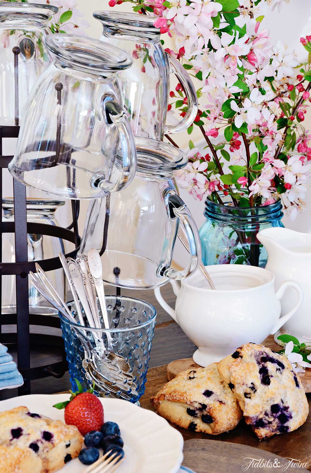 Closeup of bottle drying rack holding clear coffee mugs next to vase of cherry blossoms and plate of scones