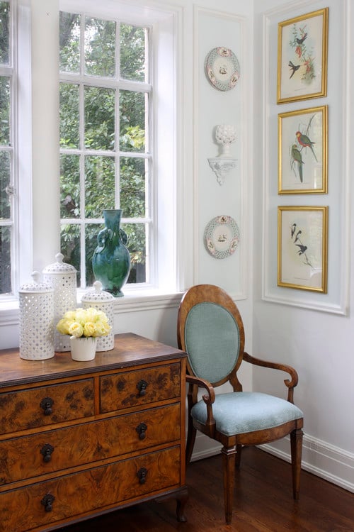 Corner of room with a small chest of drawers and a blue French chair and gold framed bird prints on the wall