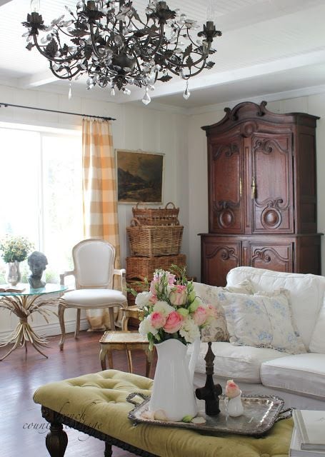 A French style living room with white slipcovered sofa and light green tufted ottoman and baskets stacked in corner