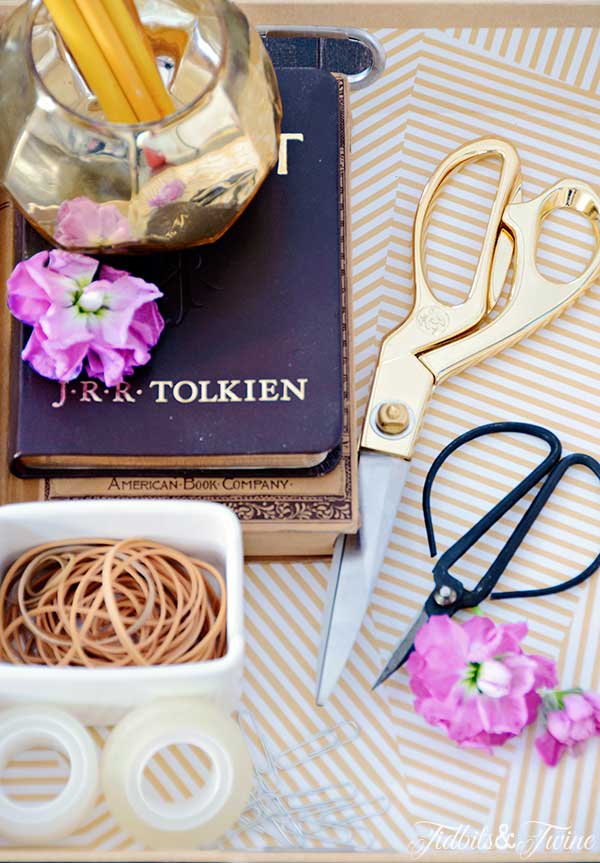 tray holding gold scissors and antique scissors with a gold vase holding pencils some purple flowers and white container of rubber bands