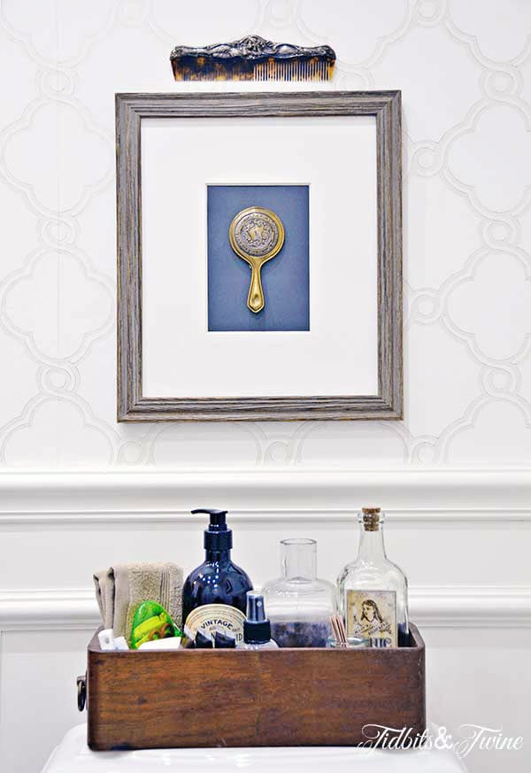 Framed silver mirror and comb hanging on wall above toilet with sewing drawer holding essentials