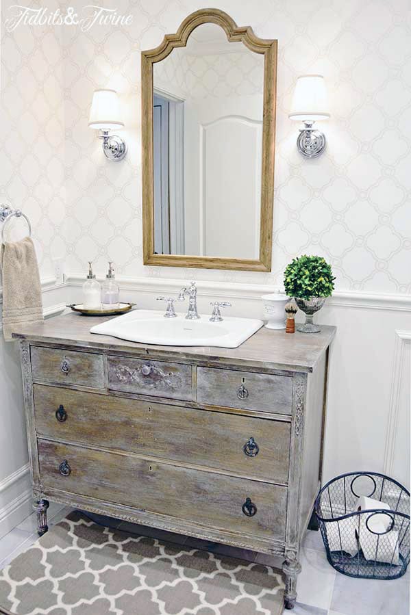 sconces with shades flank a wooden mirror in bathroom with dresser vanity wallpaper wainscotting and beadboard ceiling