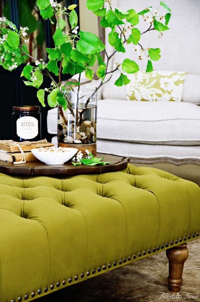 Green tufted ottoman with vase of green leaves books and candle on top