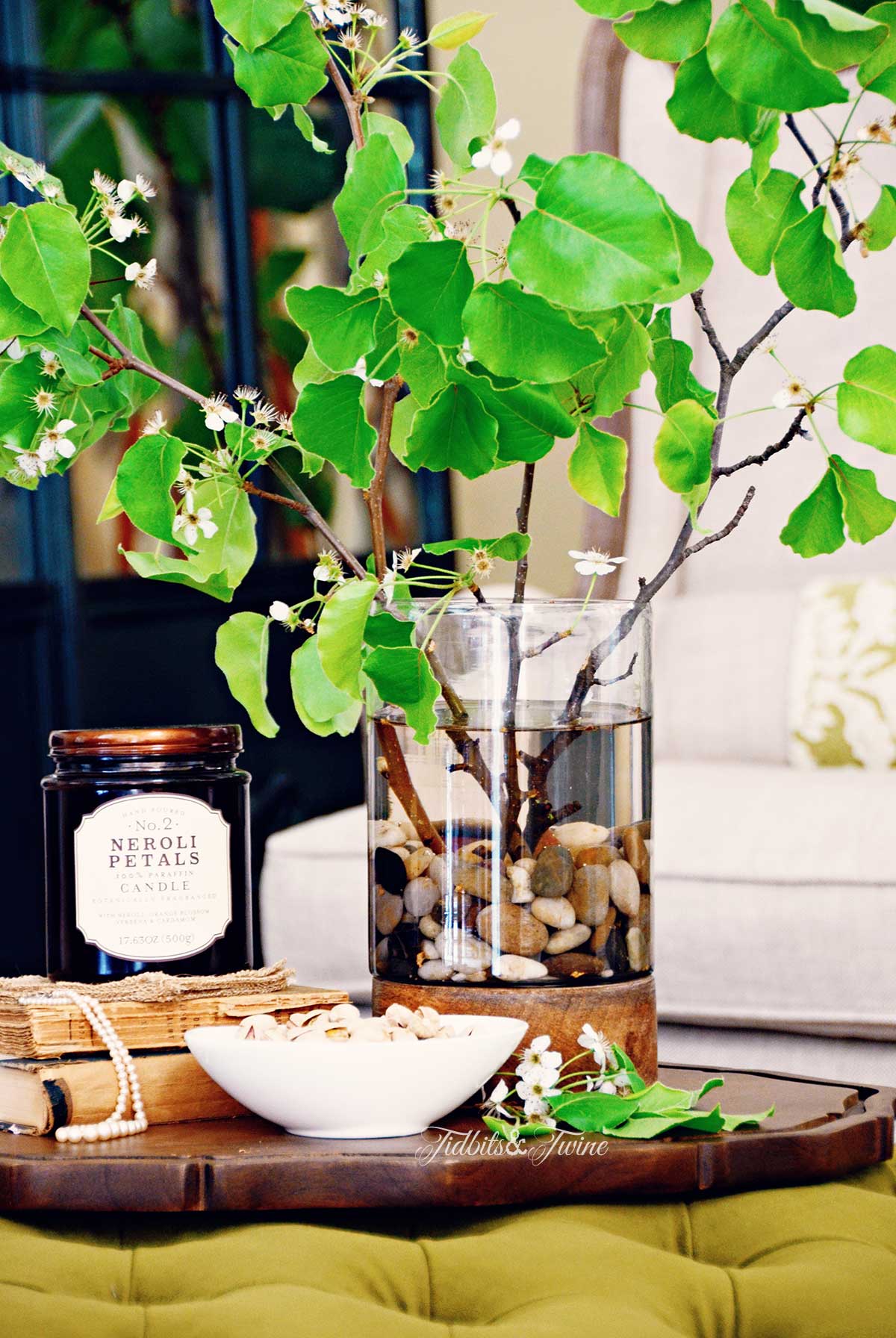 Closeup of green tufted ottoman with wood tray on top with large vase of leaves books and a candle