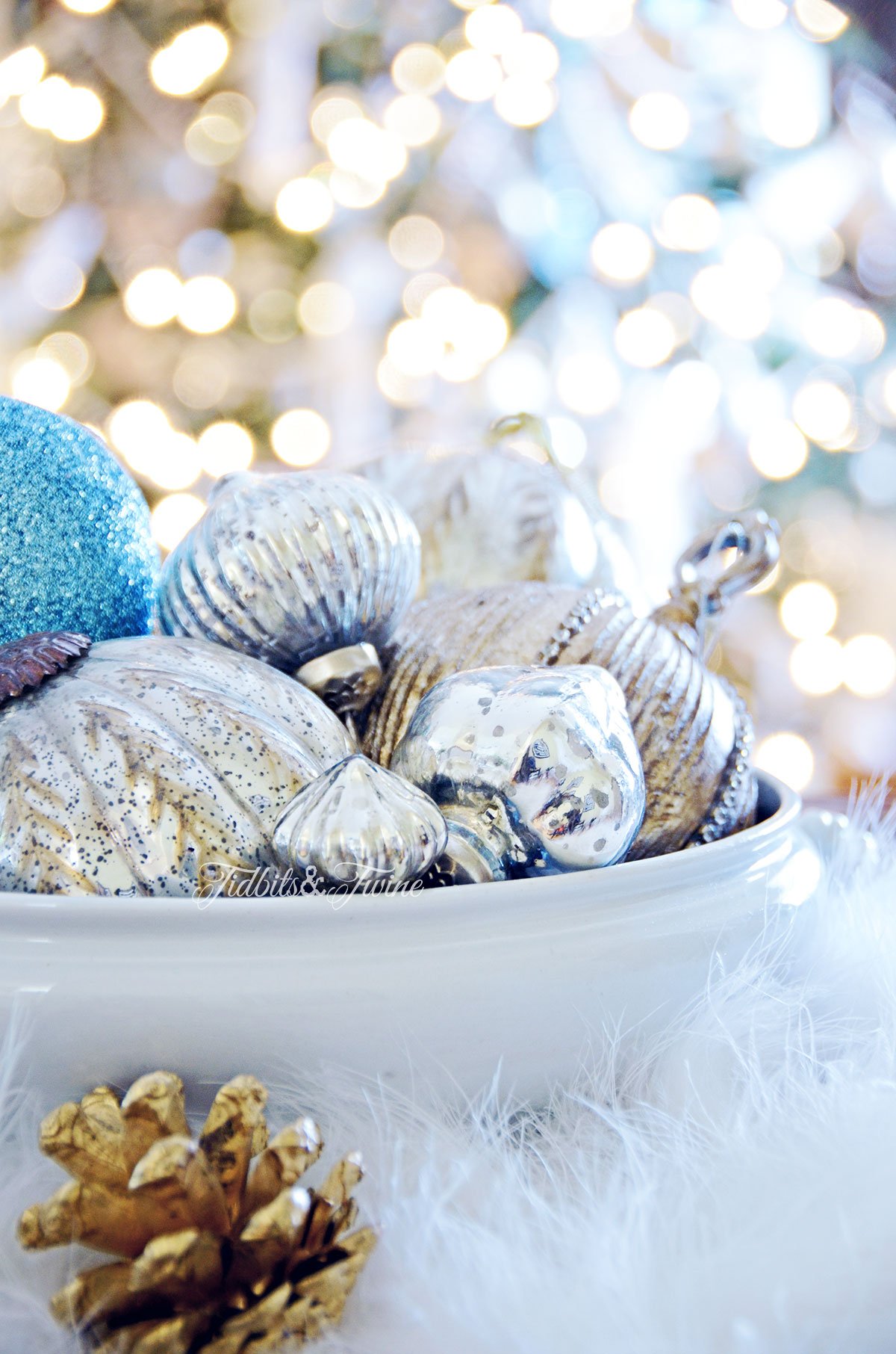 White ironstone bowl full of silver and blue ornaments with Christmas tree in the background