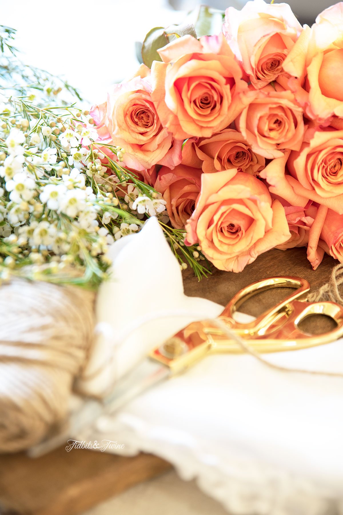 bouquet of peach roses lying on wood table next to gold scissors and white wax flowers