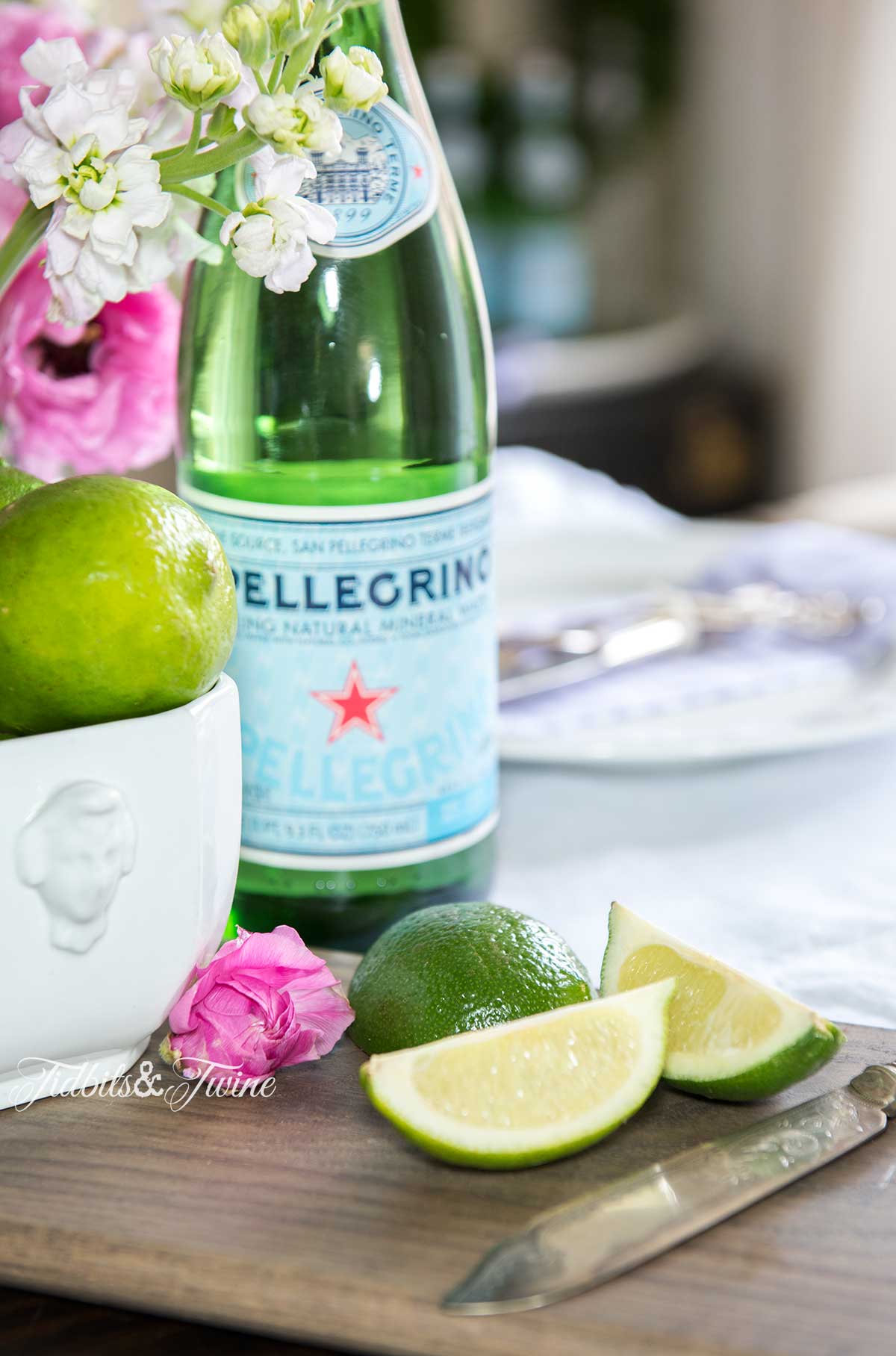 closeup of cut limes on board next to ironstone bowl of limes with pellagrino and pink flowers