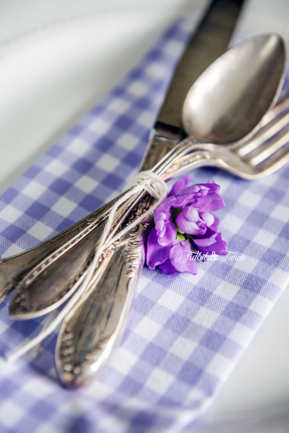 closeup of vintage flatware set tied together with twine and a purple flower on top of a purple checked napkin