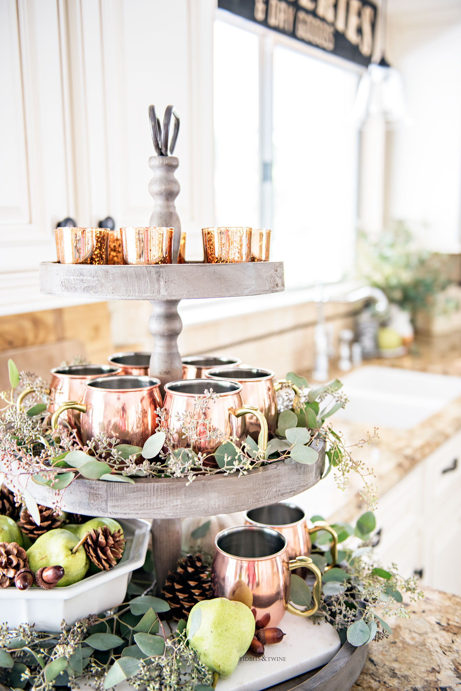 three tier wooden display holding moscow mule mugs and eucalyptus and a white bowl holding pears