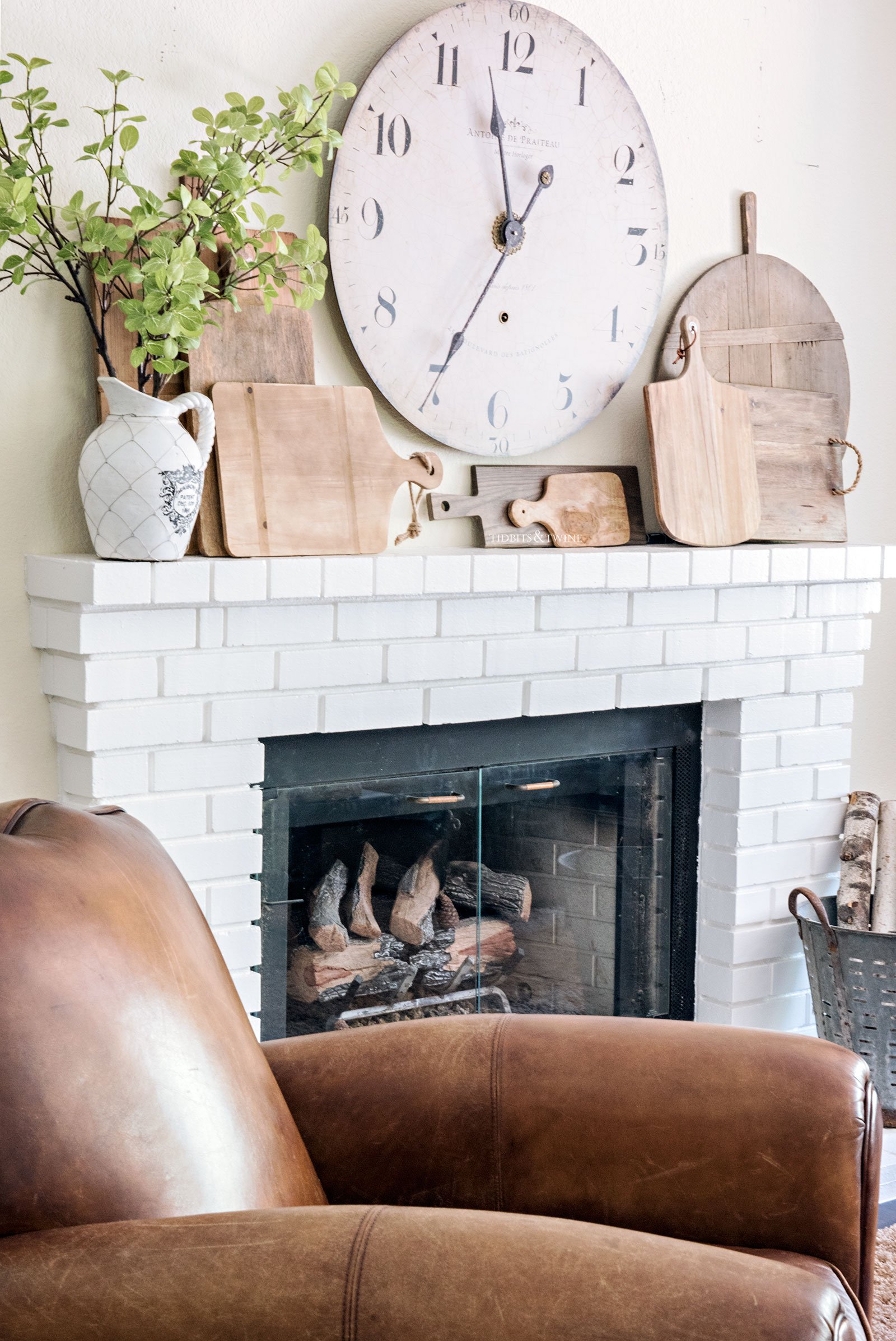 clock above a fireplace mantel decorated with a collection of breadboards and greenery