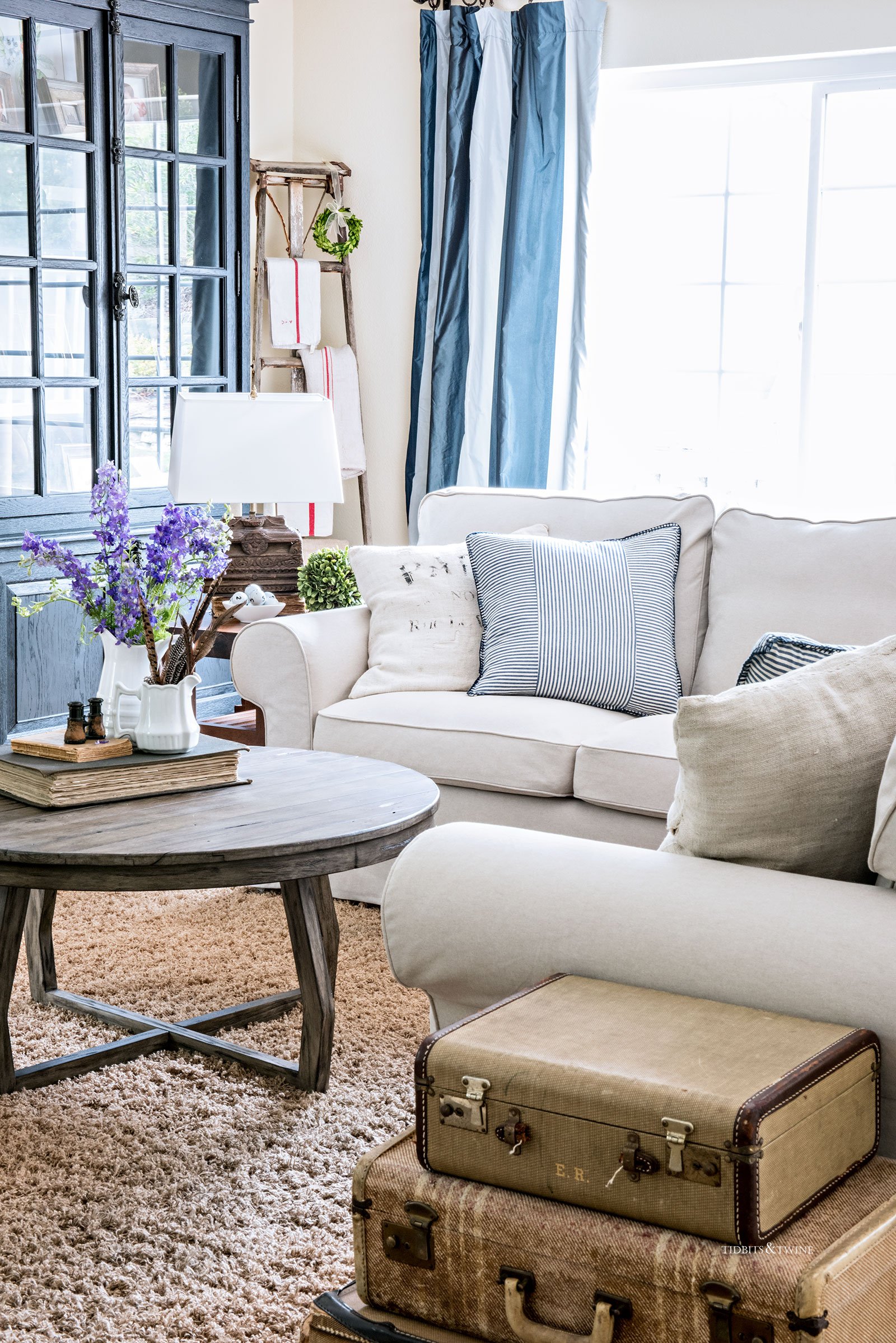 slipcovered white sectional with round coffee table blue striped curtains suitcase side table and spring decor