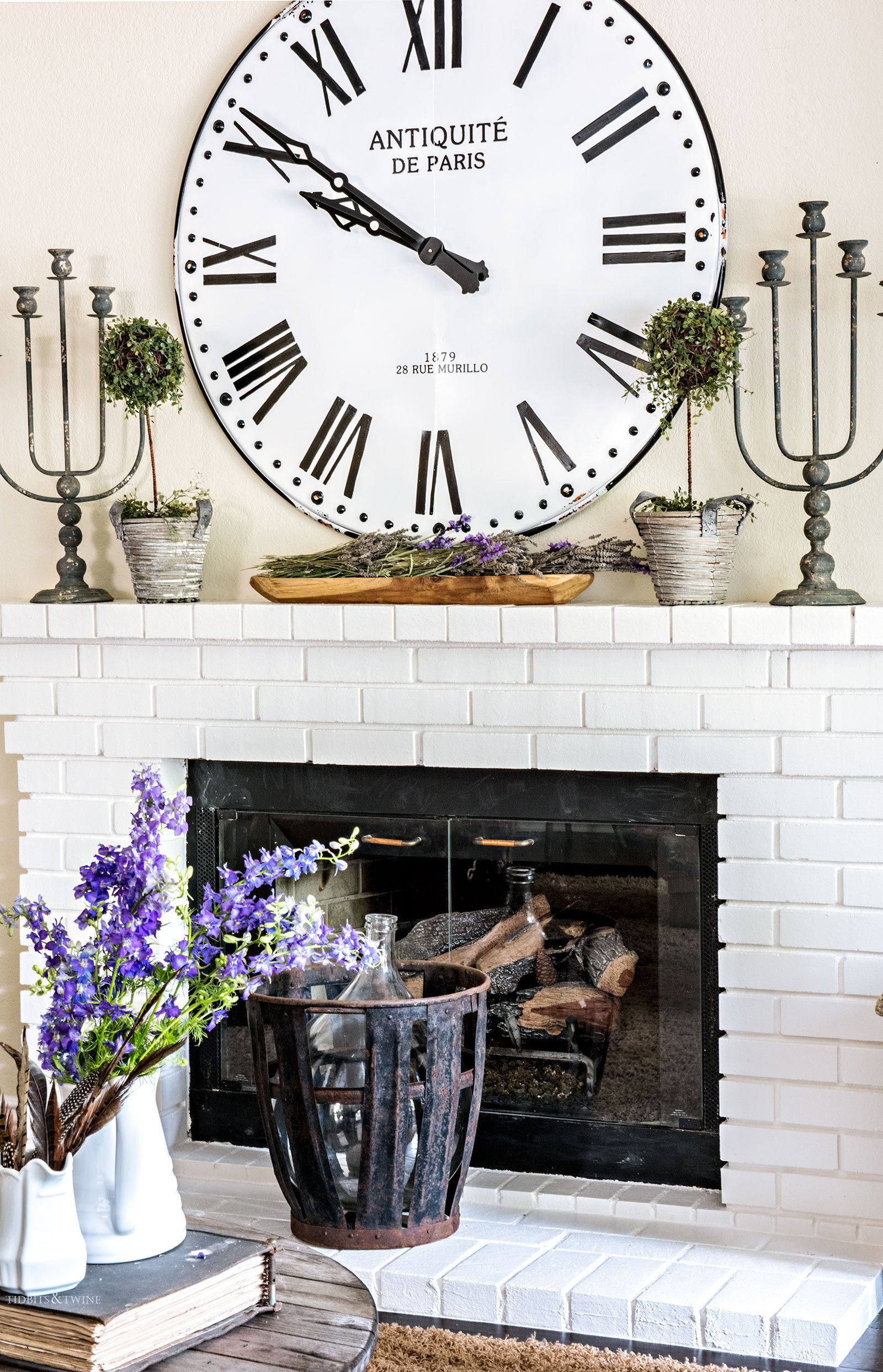 Oversized clock above a white brick fireplace with candlesticks and antique demijohn