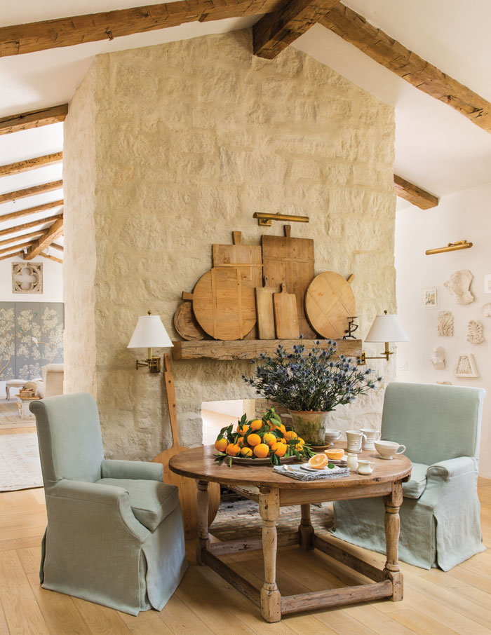 rustic european home with stone fireplace with beam mantel holding bread boards and small table in front with two blue chairs