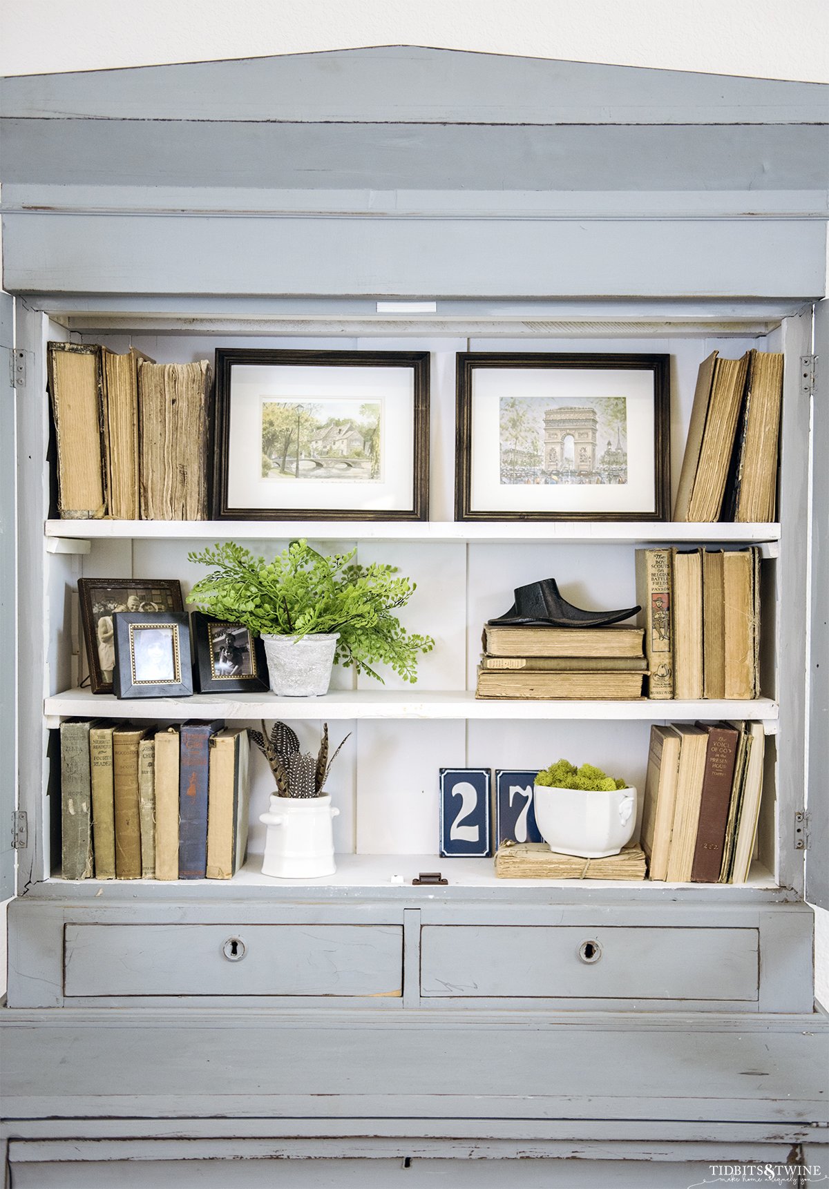 french blue cabinet styled with antique books and white ironstone and two pieces of art on the top shelf