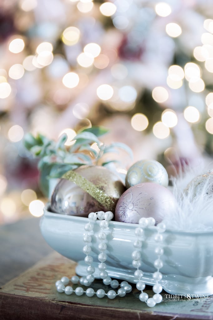 Closeup of white ironstone filled with pink Christmas ornaments and tree in background