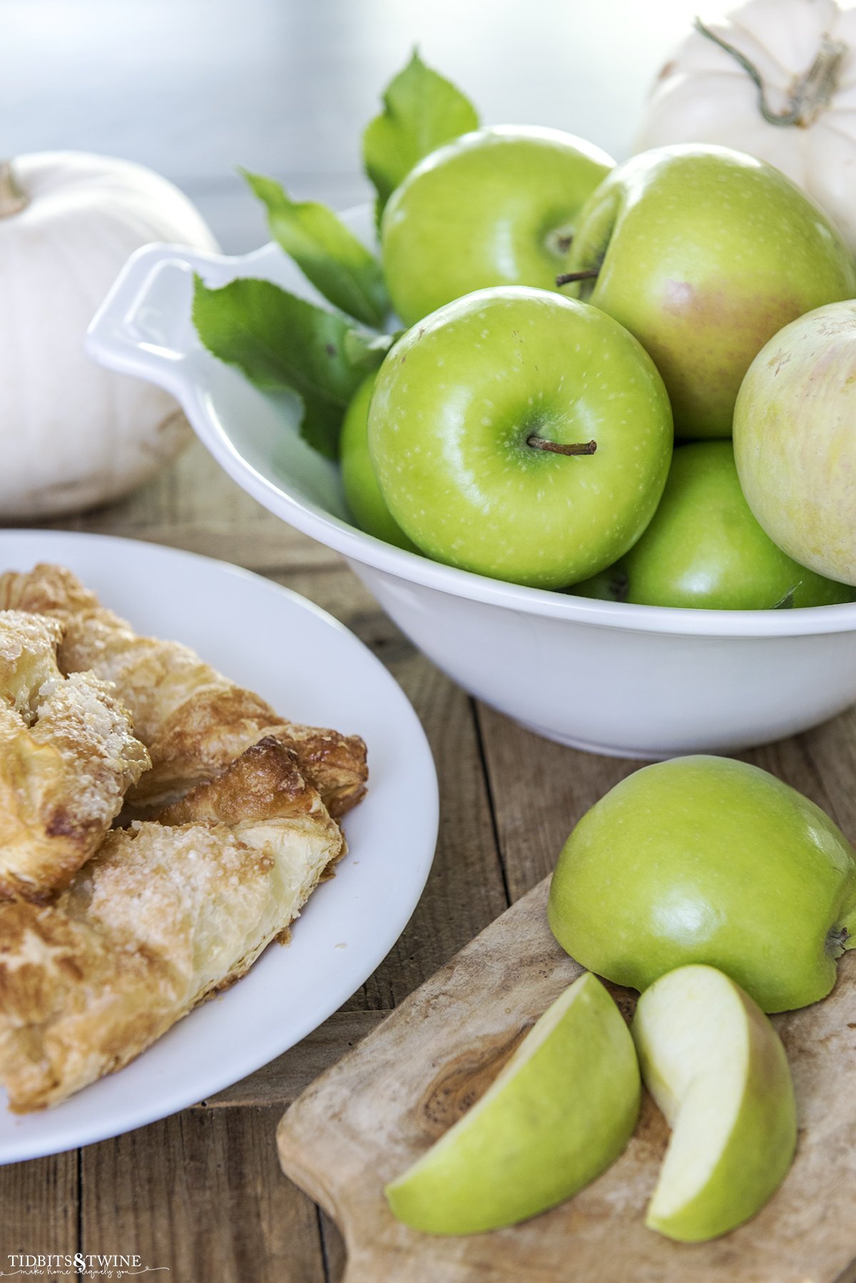 white bowl full of green apples with apple turnover on a plate and a sliced apple on cutting board