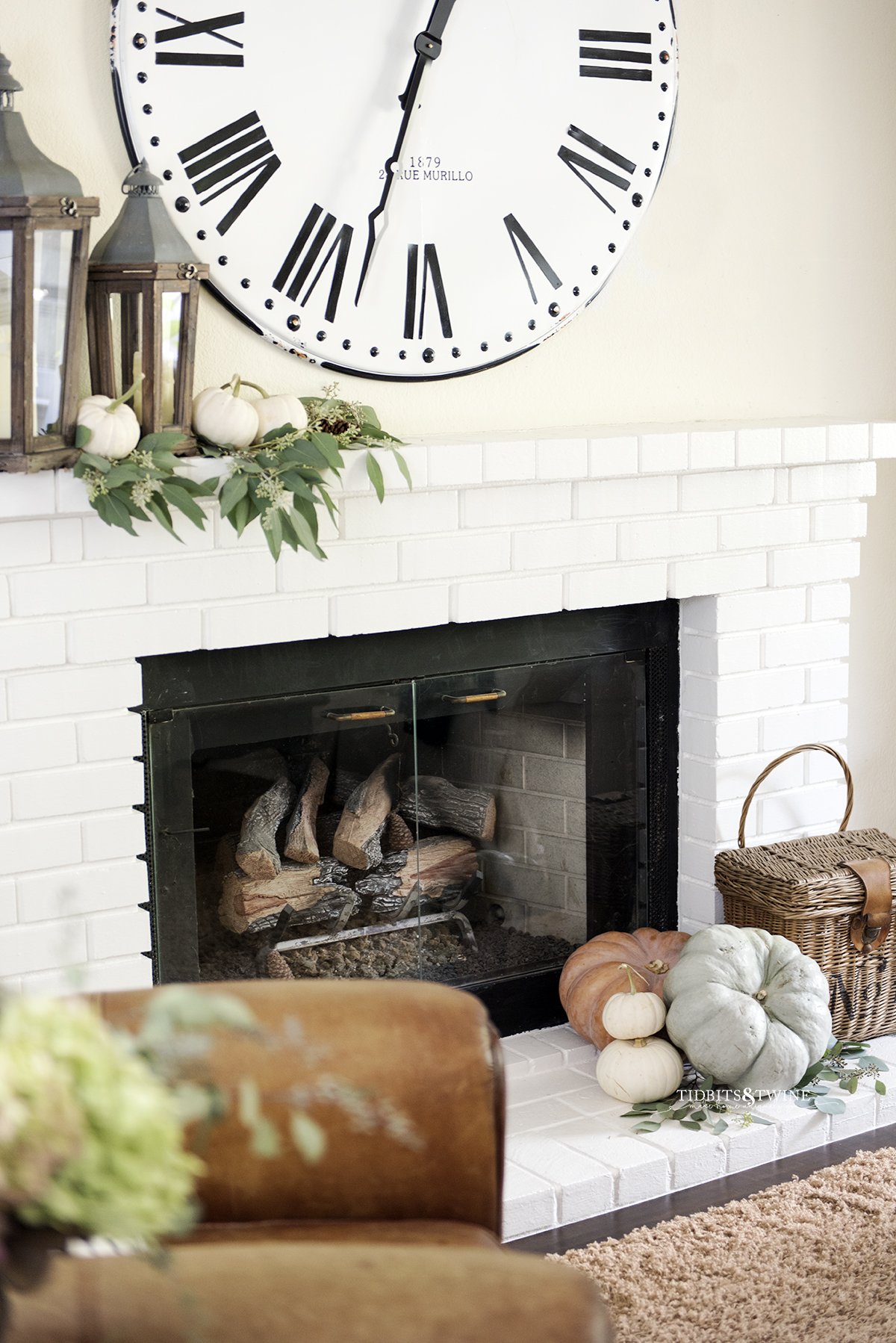 white brick fireplace mantel with large french clock above decorated for fall with pumpkins and eucalyptus