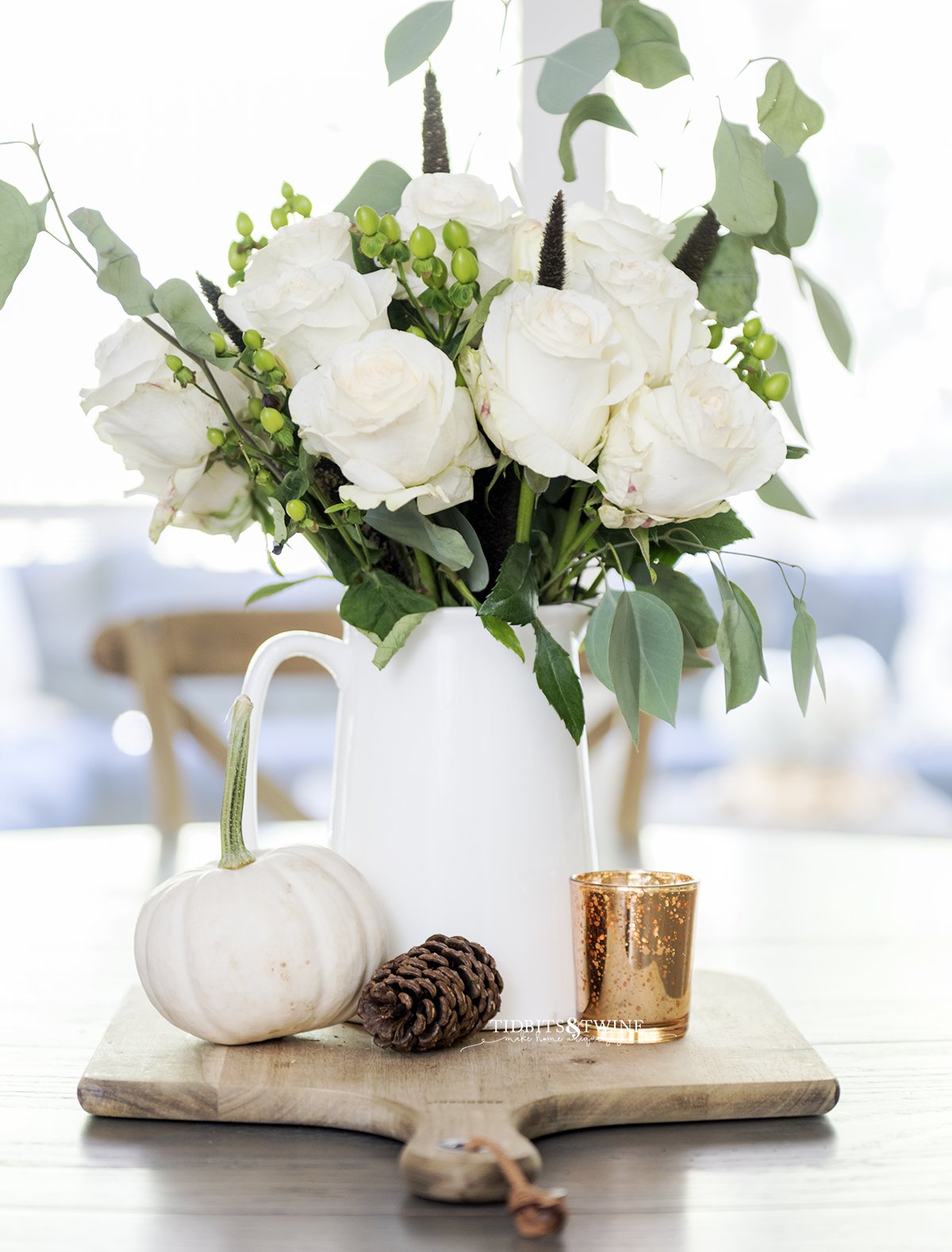 Table centerpiece for Fall with white pitcher holding white roses on a bread board with pinecone and pumpkin