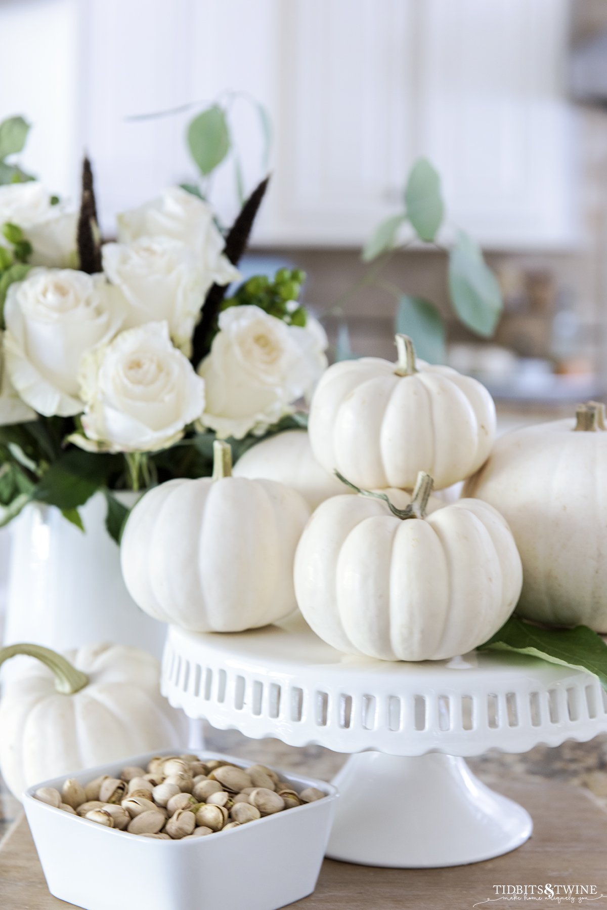 Small white pumpkins on a cake stand with a pitcher of white roses behind and a bowl of pistachios below