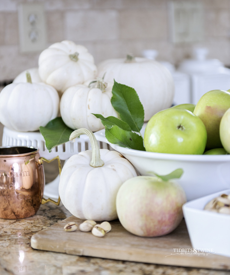 Fall vignette of green apples in a white bowl with white pumpkins on a cake stand, pistachios and a copper mug
