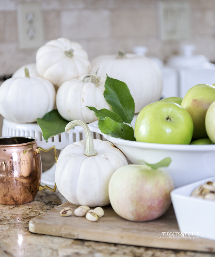 kitchen vignette with white bowl holding green apples and white pumpkins on a cake stand behind