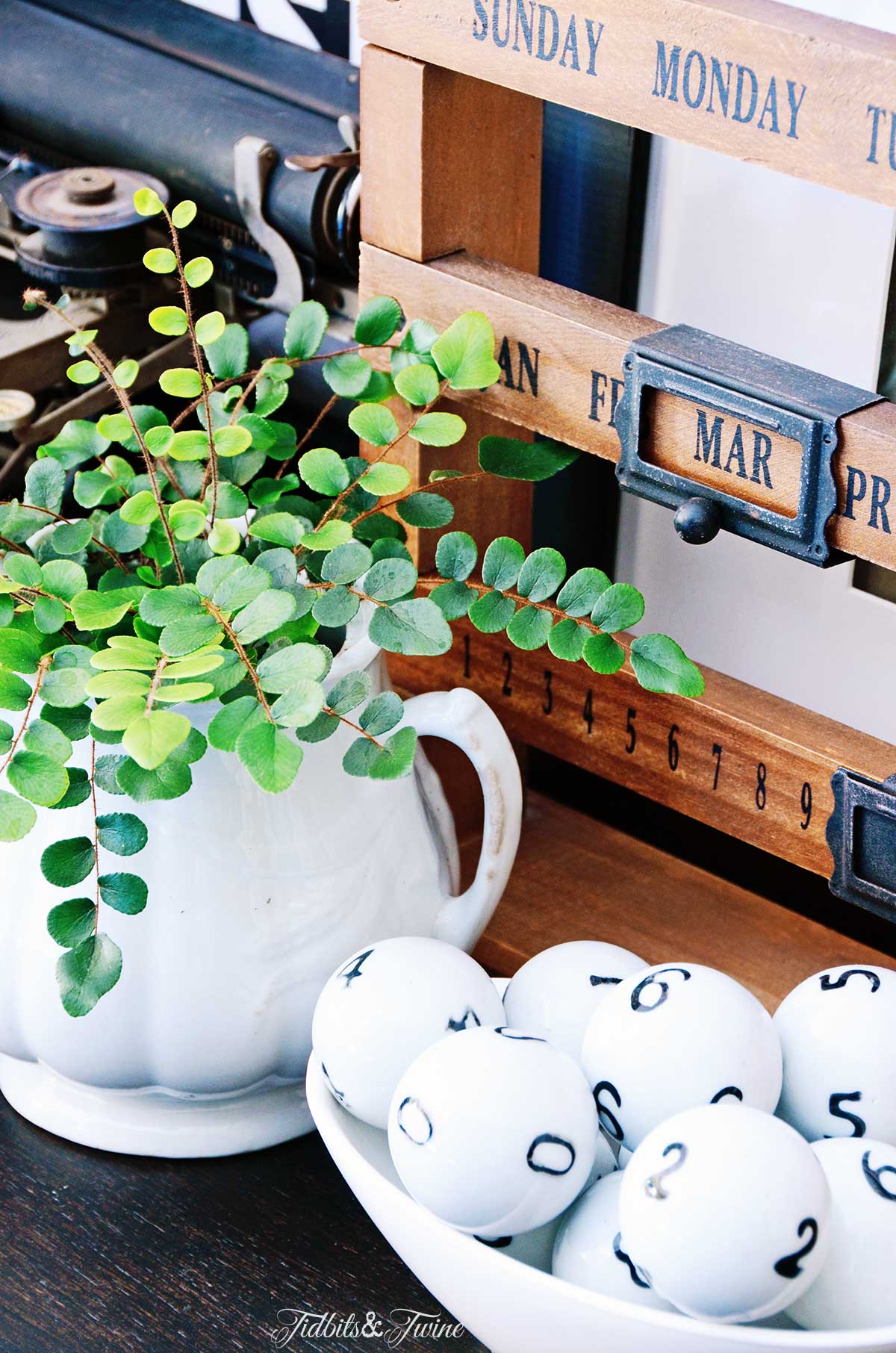 A wooden desk calendar next to antique white ironstone holding a button fern and glass numbered balls