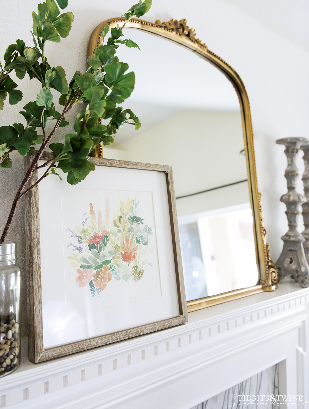 Closeup of french fireplace mantel with gold mirror and art styled on top with a vase of fake ginkgo stems