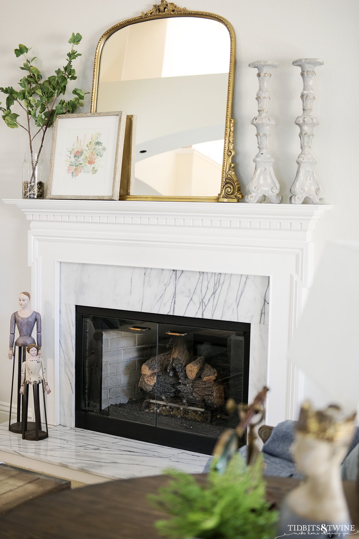 French fireplace mantel with gold mirror candlesticks and artwork on top with vase holding fake ginkgo stems
