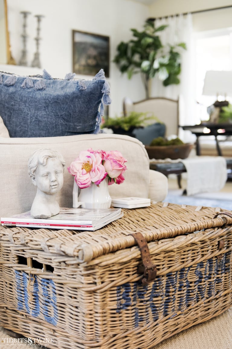 lidded french antique basket used as a side table next to sofa with small white vase of pink flowers on top