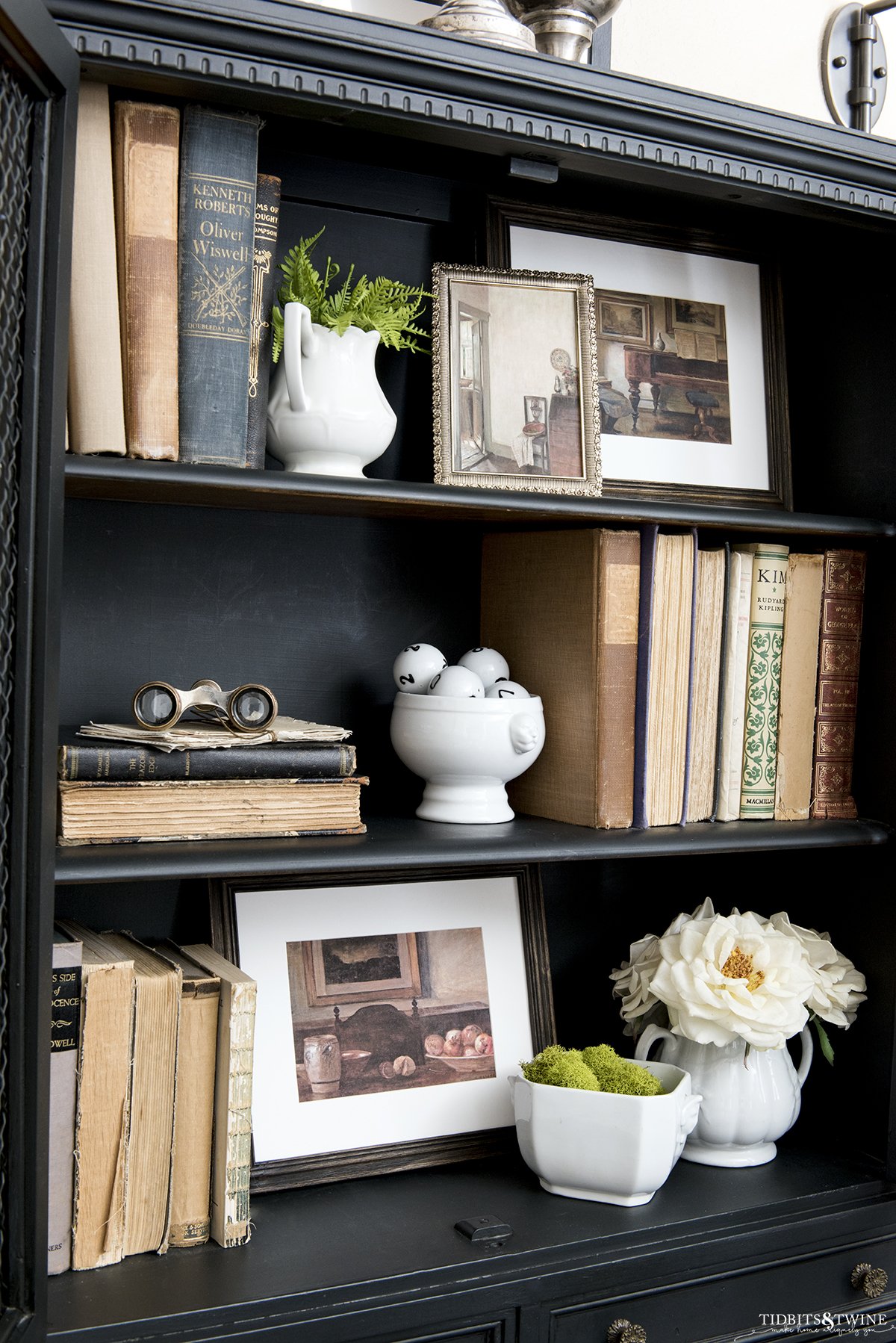 Black bookshelf styled with antique books and framed artwork and a vase of white roses