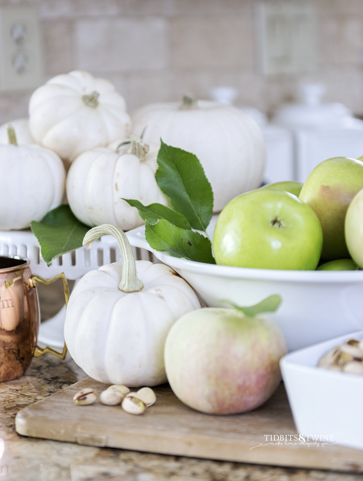 Fall vignette of green apples in a white bowl with white pumpkins on a cake stand, pistachios and a copper mug