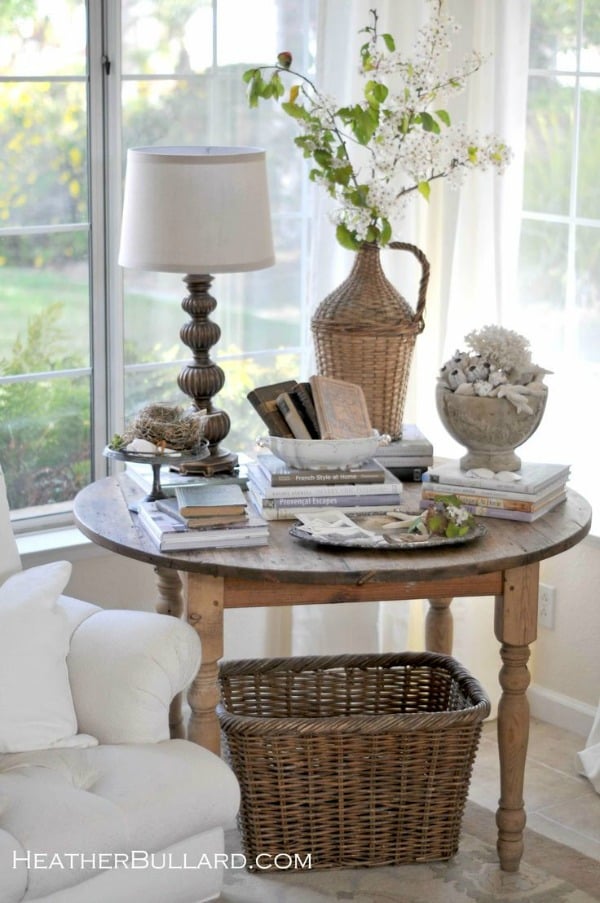 antique wooden table in corner of living room windows with books baskets and shells on top and a large basket underneath