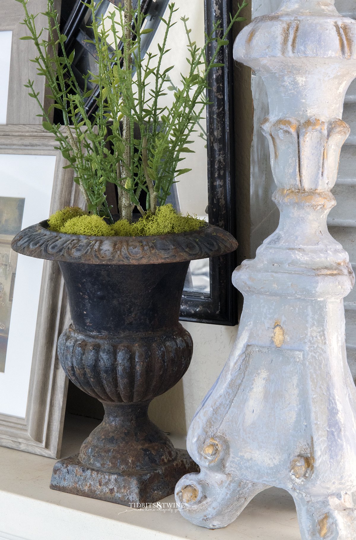 Closeup of antique french metal urn with myrtle topiary inside and green moss at base next to a french candlestick on a mantel