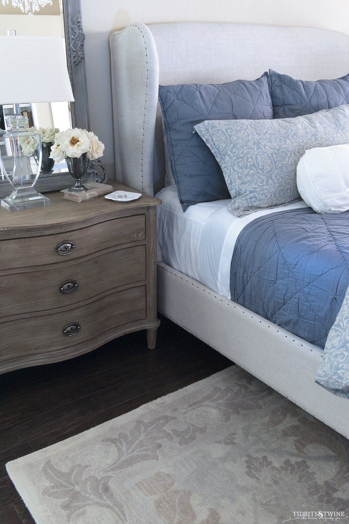 wingback linen headboard with blue bedding next to wood nightstand with crystal lamp and french mirror