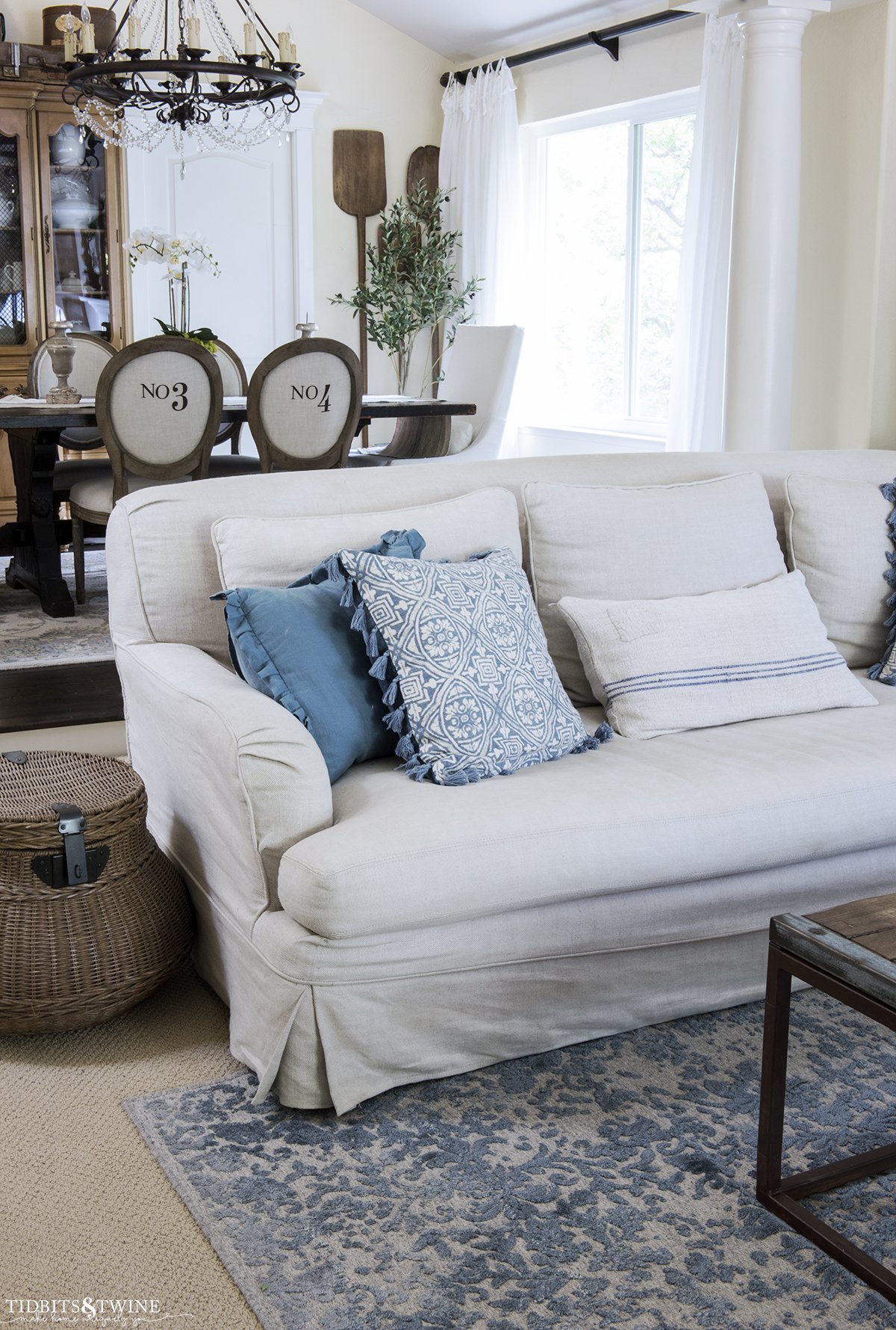 slipcovered living room sofa with blue pillows and blue rug underneath looking into french dining room with numbered chairs