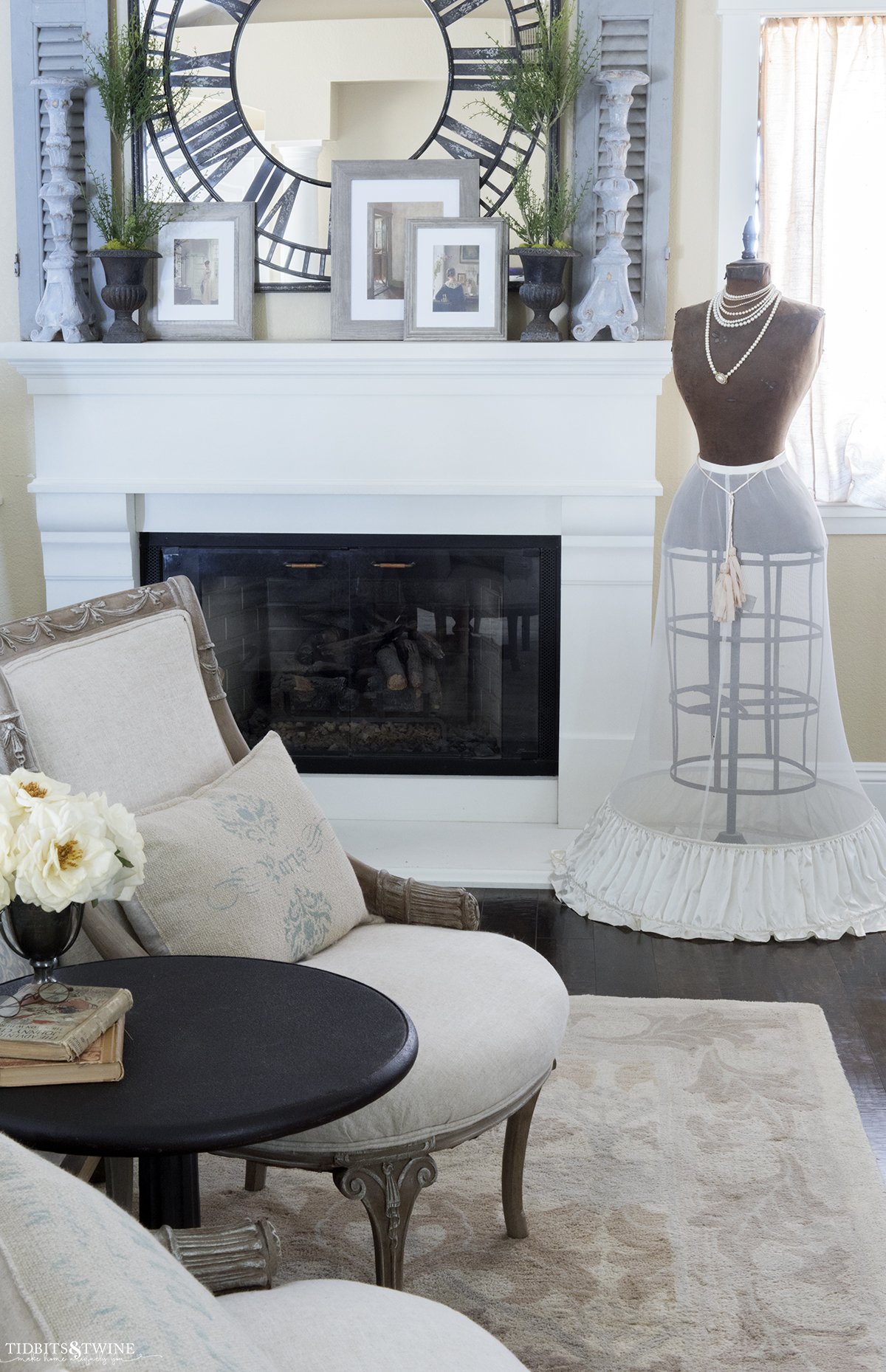 french seating area at end of bed looking toward white cast stone fireplace mantel with mirror and candlesticks above and antique dress form on side