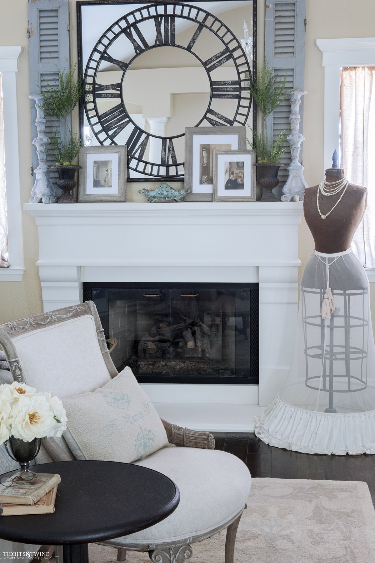 french seating area at end of bed looking toward white cast stone fireplace mantel with mirror and candlesticks above and antique dress form on side