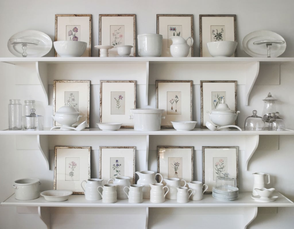 three white shelves holding a white ironstone collection with framed antique botanicals in leaning against the wall