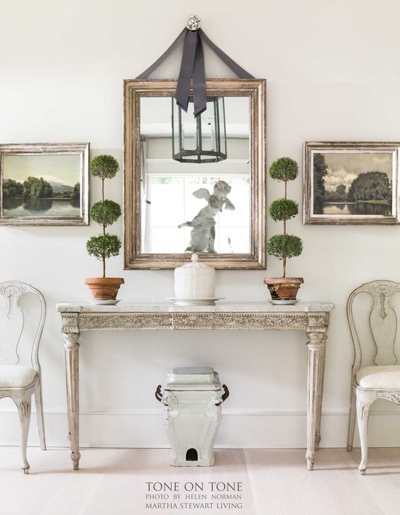 gustavian console table with gold mirror above hanging from gray ribbon and two triple ball myrtle topiaries on either side