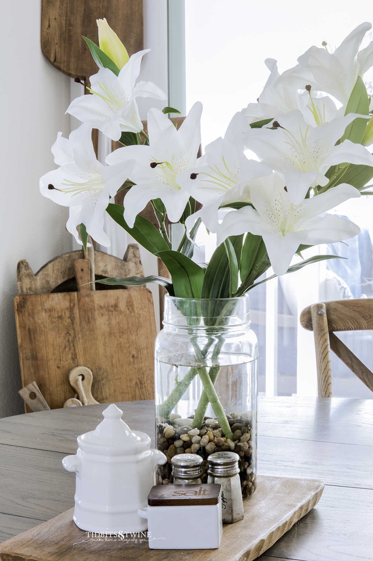 artificial white lilies in an antique pickling jar on a kitchen table next to sugar and salt and breadboard collection in the background