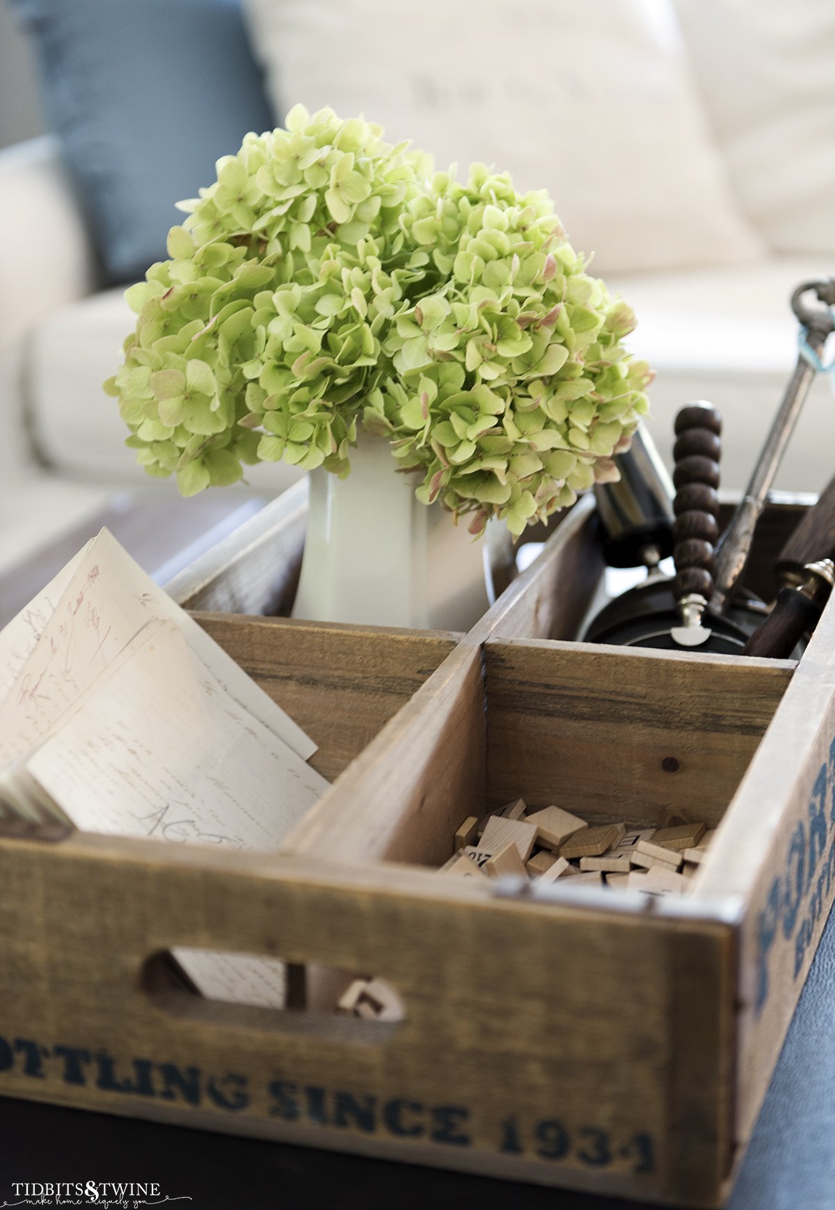 white ironstone vase filled with green hydrangea that are drying in a wooden box with magnifying glasses and scrabble tiles on coffee table