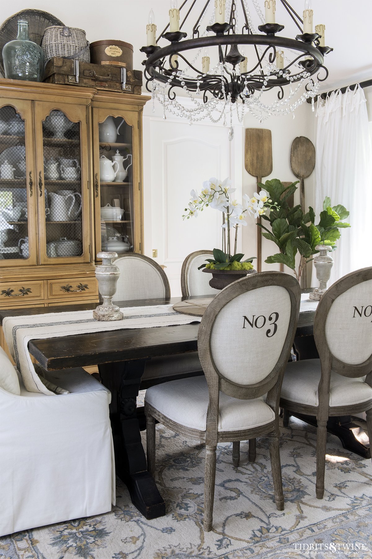 round french linen chairs with numbers on the back in dining room with white orchid on the table and vintage bread paddles hanging on the wall