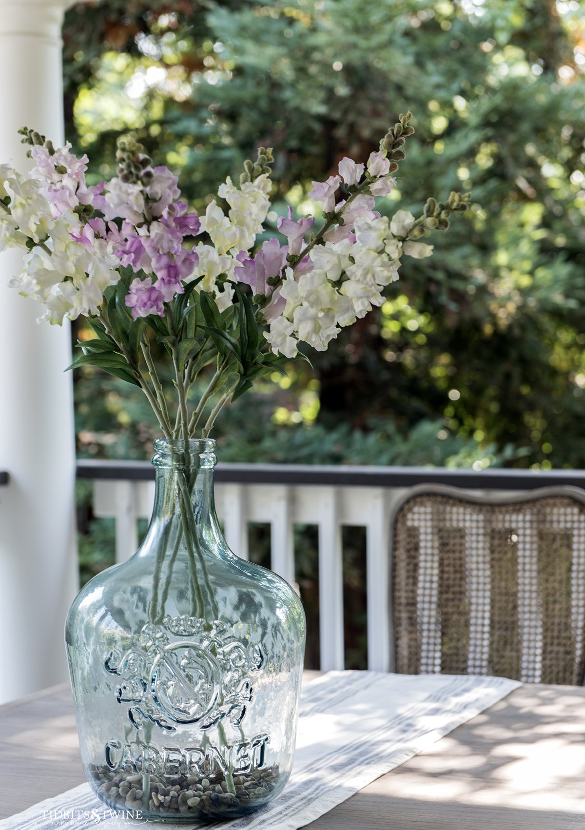 blue glass demijohn filled with white and purple fake snapdragons on a table outside on a porch