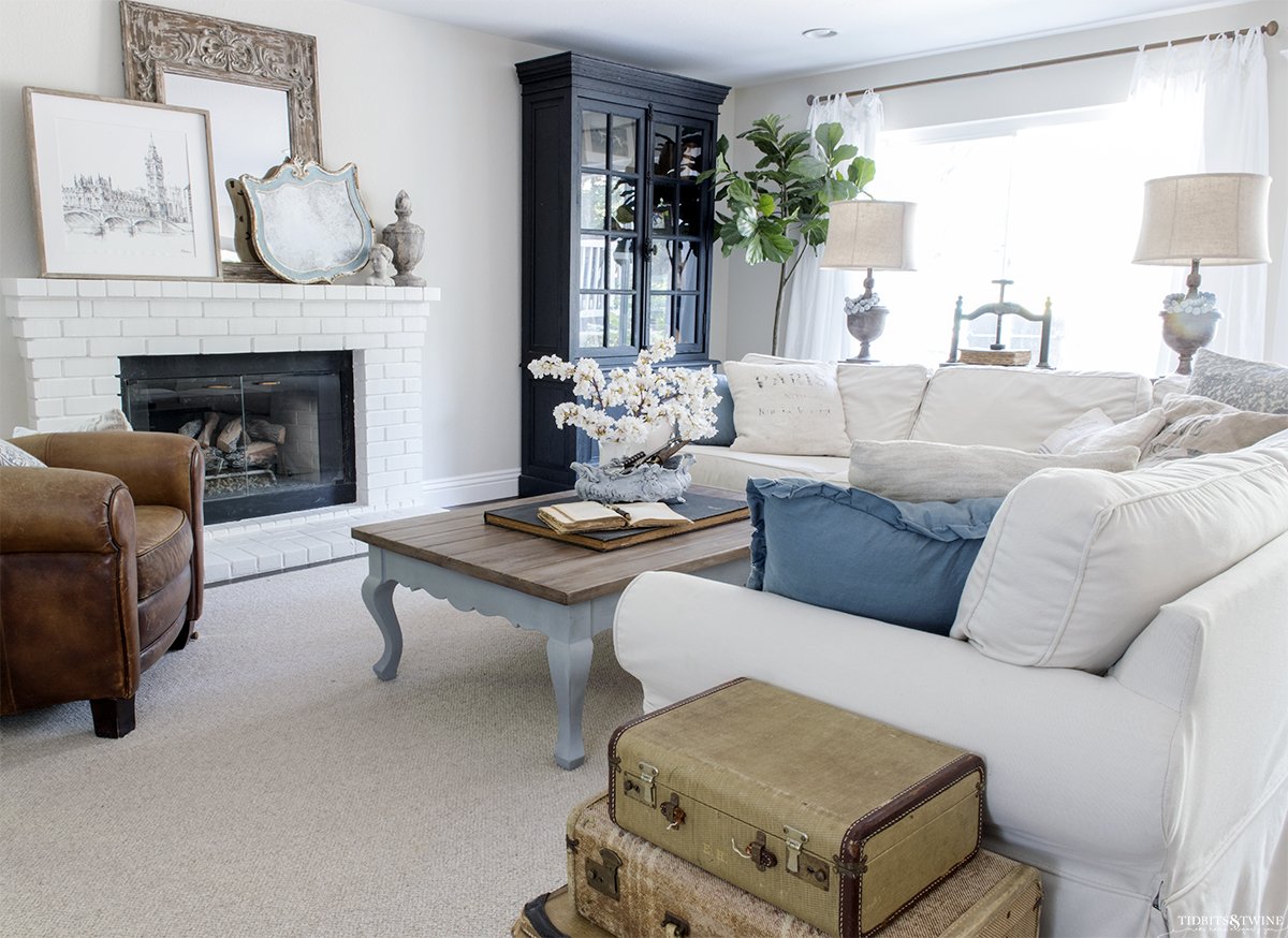 white slipcovered sectional with blue pillows and french blue coffee table white brick fireplace and black french cabinet