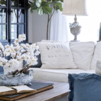 living room with white slipcovered sectional and blue throw pillows french coffee table with cherry blossoms in a vase on top