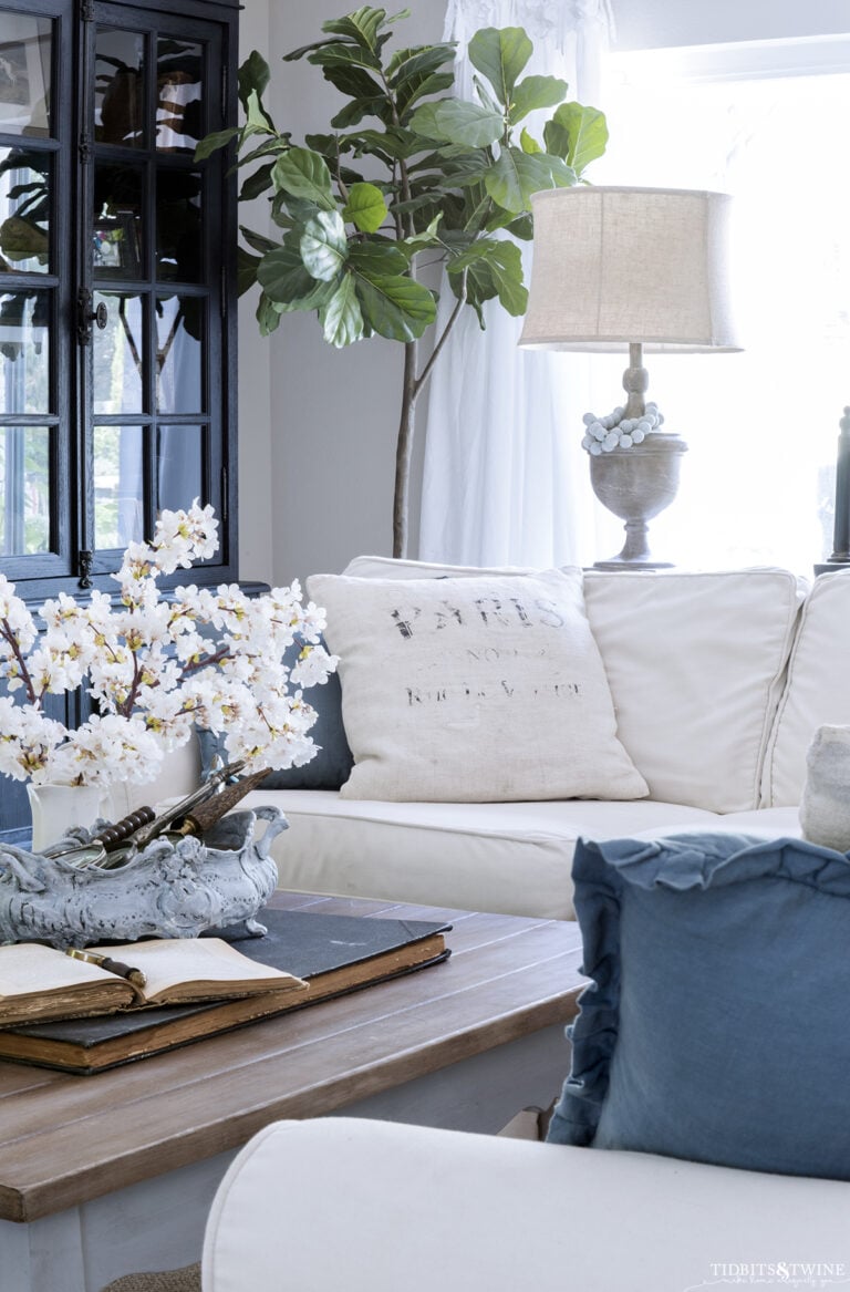 living room with white slipcovered sectional and blue throw pillows french coffee table with cherry blossoms in a vase on top