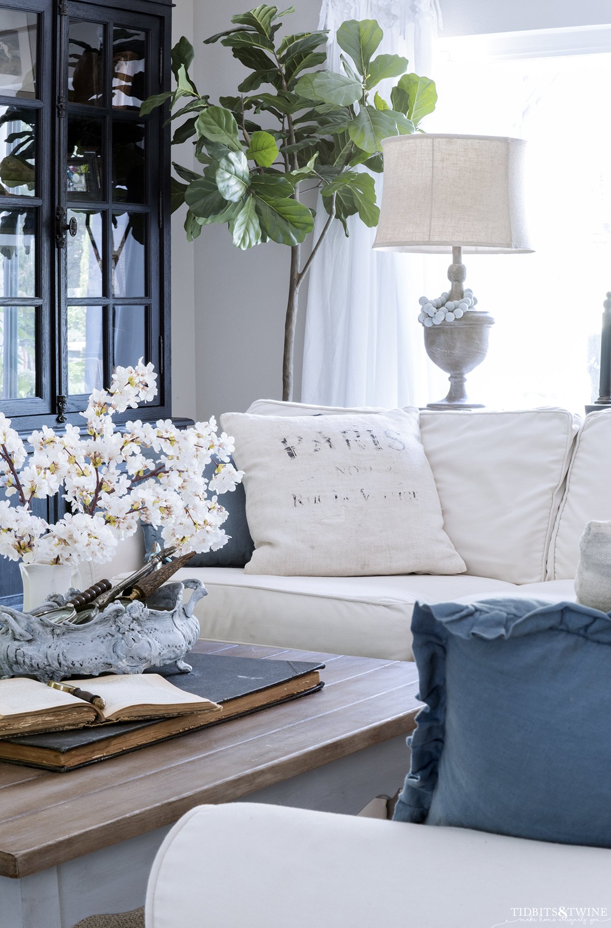 living room with white slipcovered sectional and blue throw pillows french coffee table with cherry blossoms in a vase on top