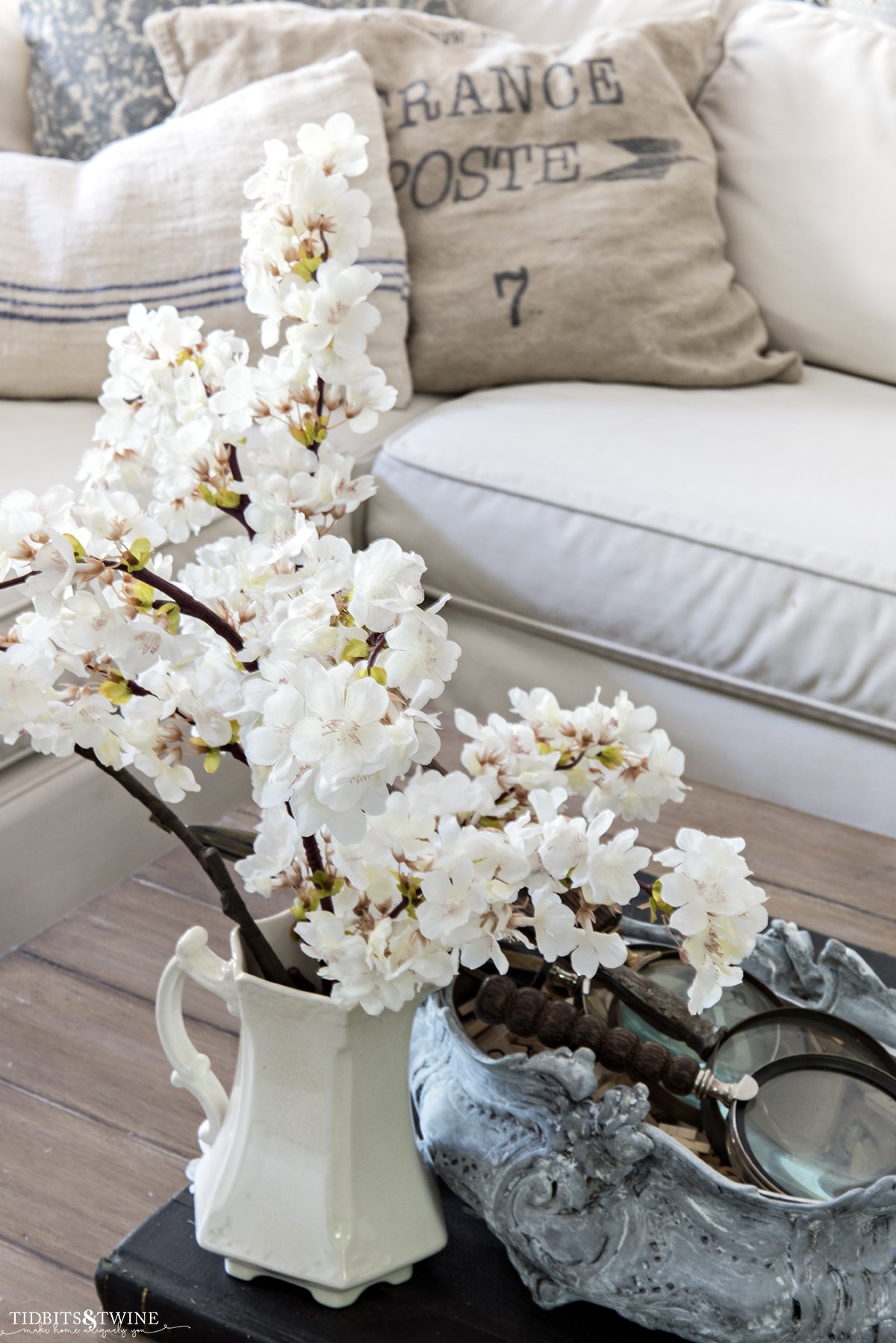 cherry blossom stems in an ironstone pitcher on coffee table with sectional sofa in background with french pillows